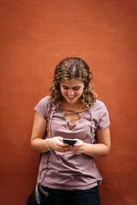 Young woman using phone while standing against orange wall