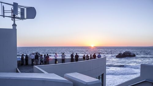 People standing by sea against clear sky during sunset