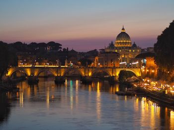 Illuminated bridge over river by buildings against sky during sunset