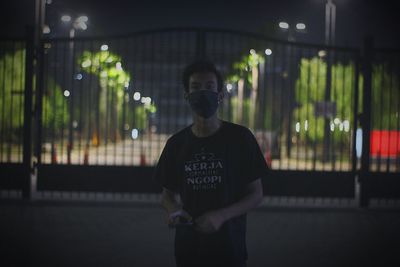 Portrait of young man standing against illuminated wall at night