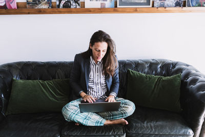 High angle of young female entrepreneur in formal blazer and pajama pants sitting on sofa and communicating with partner via video call on laptop while working remotely from home