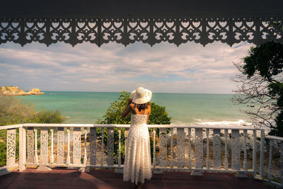 Young woman with white dress and hat are relaxing at balcony in bungalow on the beach in summer 