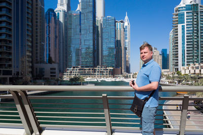 Portrait of young man standing by railing against buildings in city