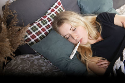 Ill young blonde woman lying in bed and holding a thermometer in her mouth.