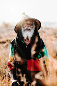 Cheerful old ethnic rastafari with dreadlocks looking at the camera in standing in a dry meadow in the nature