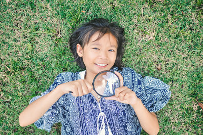 High angle portrait of girl holding magnifying glass while lying on grassy field