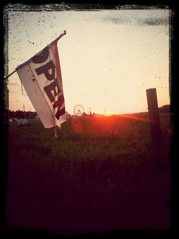transfer print, sky, sunset, guidance, sunlight, auto post production filter, flag, text, communication, outdoors, no people, western script, identity, arrow symbol, transportation, sun, directional sign, patriotism, field, day