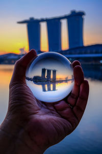 Close-up of hand holding crystal ball against sky during sunset