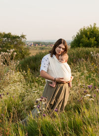 Woman with baby carrying in fabric while standing on field against sky