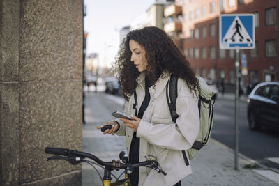 Female construction freelancer pairing bluetooth headphones with smart phone while standing by bicycle near wall at stre