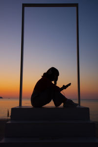 Silhouette of man sitting on sea against sky during sunset