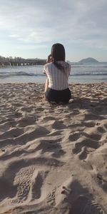 Rear view of young woman looking at sea while sitting on beach against sky