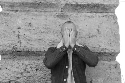 Man covering face while standing against stone wall 