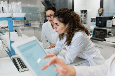 Young female pharmacy researchers gathering at table with laptop with diagrams and discussing results of research while working together in scientific lab