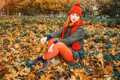 Autumn portrait of cute girl in orange warm knitted hat and orange scarf sitting in fallen leaf