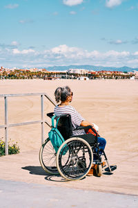 Side view of handicapped female in protective mask sitting in wheelchair with backpack and enjoying sunny summer day on sandy beach