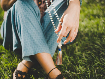 Low section of woman holding necklace while sitting on grassy field