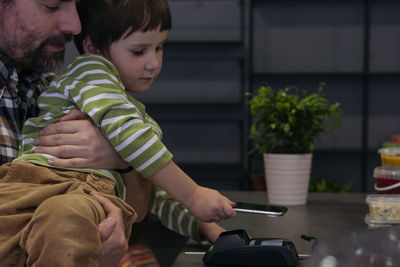 A toddler boy paying. contactless with a smartphone while his father is holding him