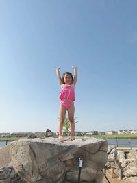 Full length of smiling girl with arms raised standing on rock against clear blue sky