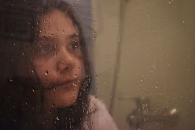 Close-up of girl looking through wet glass window in rainy season