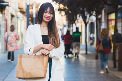 Portrait of beautiful young woman holding paper bag while standing in city