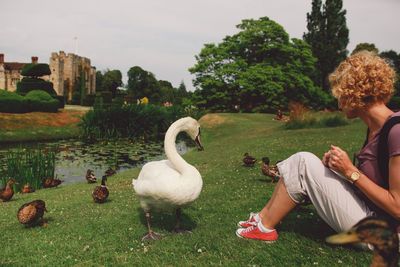 Woman sitting on grassy lakeshore with birds against sky