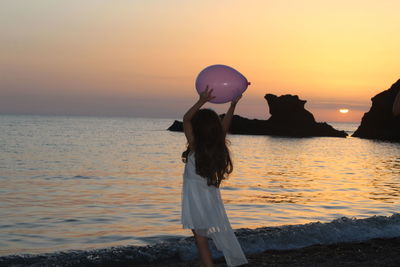 Girl holding balloon while standing by sea against sky during sunset
