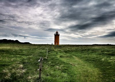 Trail on grassy field leading towards lighthouse against cloudy sky