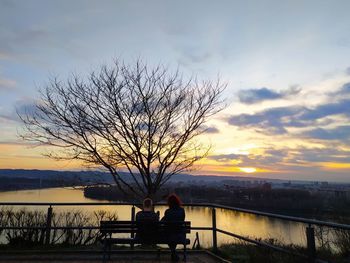 Silhouette people sitting on bench by bare tree against sky during sunset