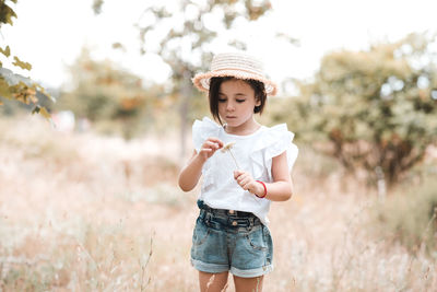 Pretty child girl 3-4 year old wearing summer clothes and hat hold flower posing in meadow closeup