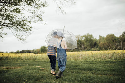 Rear view of boys walking with umbrella on grassy field against sky