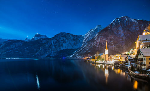 Illuminated buildings by mountains and sea against sky at dusk