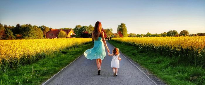 Full length rear view of mother and daughter holding hands while walking on road amidst field