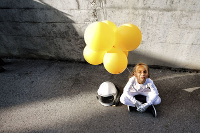 High angle view of girl holding yellow balloon sitting against wall during sunny day