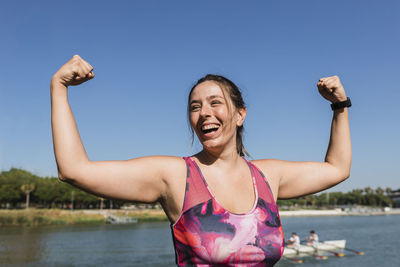 Sportswoman laughing while flexing muscles at riverbank