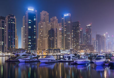 Boats moored in harbor against illuminated buildings in city at night