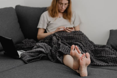 Attractive young woman working from home - female entrepreneur sitting on sofa with laptop computer and checking cell phone from comfort of home