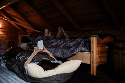 Male and female friends using mobile phones while lying in cottage during bedtime