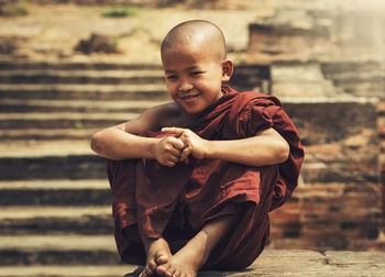 Smiling monk sitting on retaining wall against steps outside temple