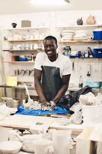 Portrait of happy young man wearing apron molding clay standing at table in art class