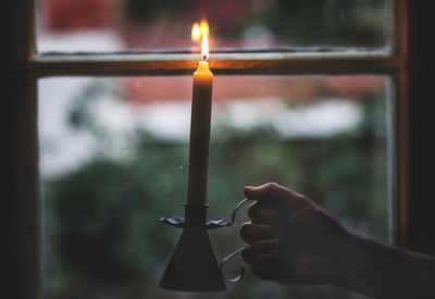 Close-up of person holding burning candle against window
