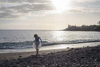 Rear view of woman standing on shore at beach against sky during sunset