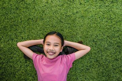 Directly above portrait of smiling girl with hands behind head lying on grassy field