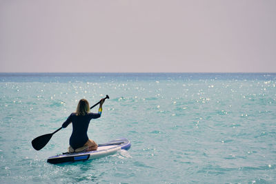 Young woman dressed in swimsuit paddling on stand up paddle board opposite turquoise colored sea