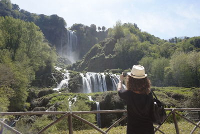 Rear view of woman photographing waterfall while standing at observation point