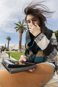 Young woman using mobile phone while sitting against sky