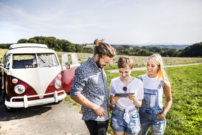 Happy friends outside van in rural landscape looking at tablet