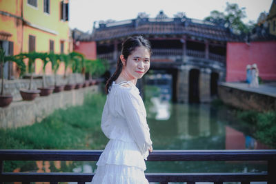 Portrait of smiling young woman standing by railing of bridge over canal
