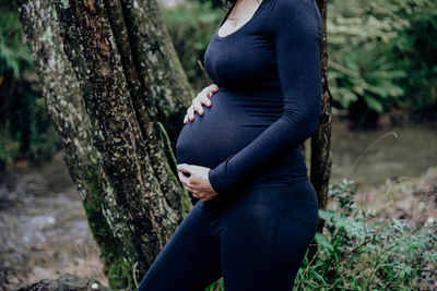 Midsection of pregnant woman standing by tree trunk