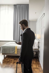 Side view of businessman using mobile phone standing with luggage in hotel room